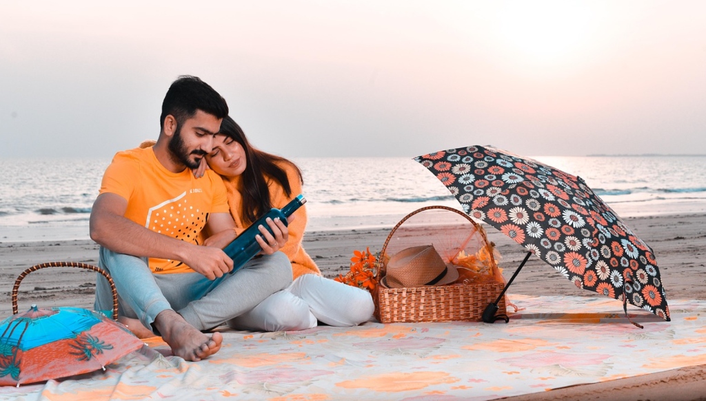 Authentic couple enjoying relaxed beach picnic with fruit basket and umbrella - wholesome dating