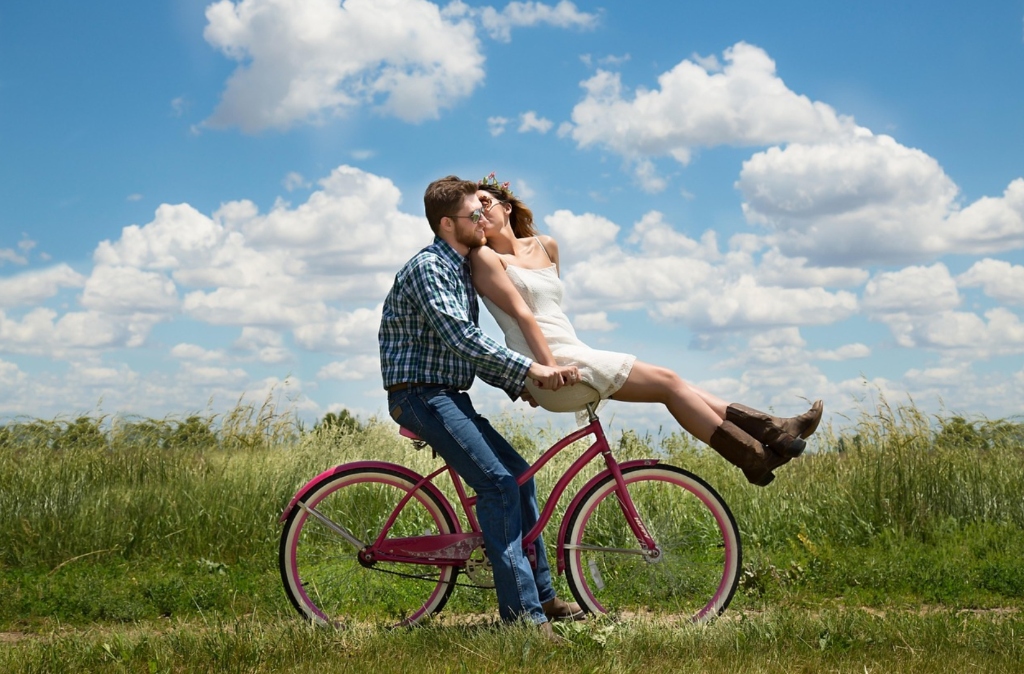 Joyful romantic couple riding bicycle together in sunlit summer countryside meadow with blue sky
