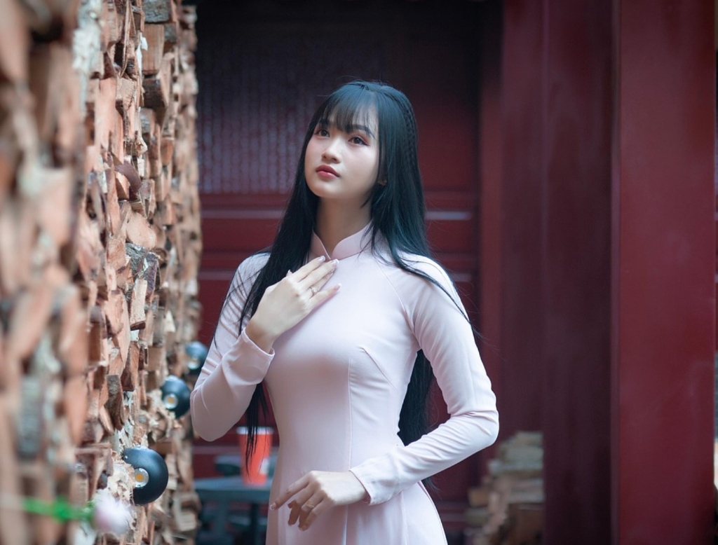 Traditional Vietnamese woman wearing elegant pink Ao Dai dress standing near firewood wall