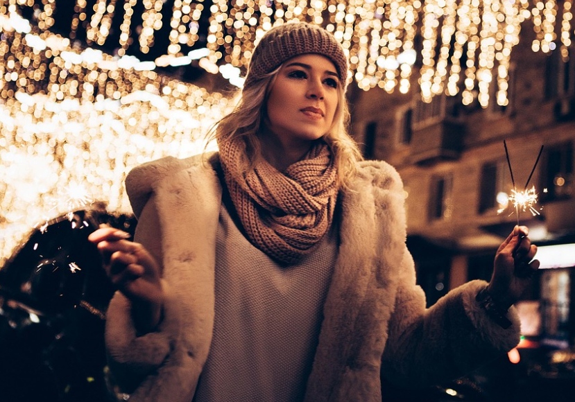 Radiant Ukrainian woman in warm knit hat and coat holding sparklers under golden fairy lights in winter city